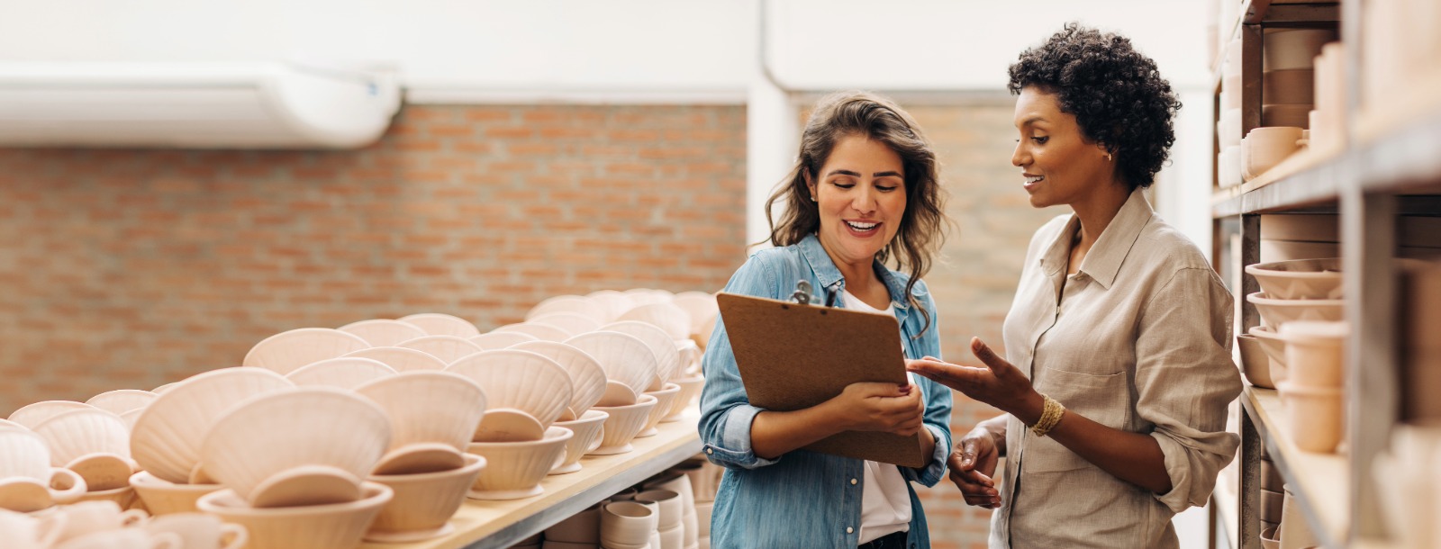 Two women in a ceramic shop talking and looking at a clipboard in the hand of the woman on the left.